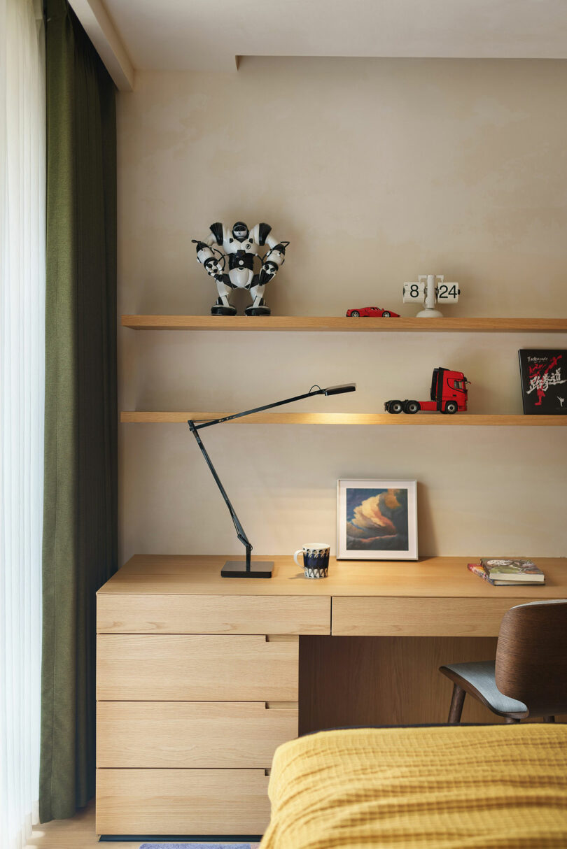 A modern wooden desk with a sleek black lamp, pencils in a holder, and a framed picture. Shelves above display decor items including a robot figure and toy trucks. A window with curtains is on the left.