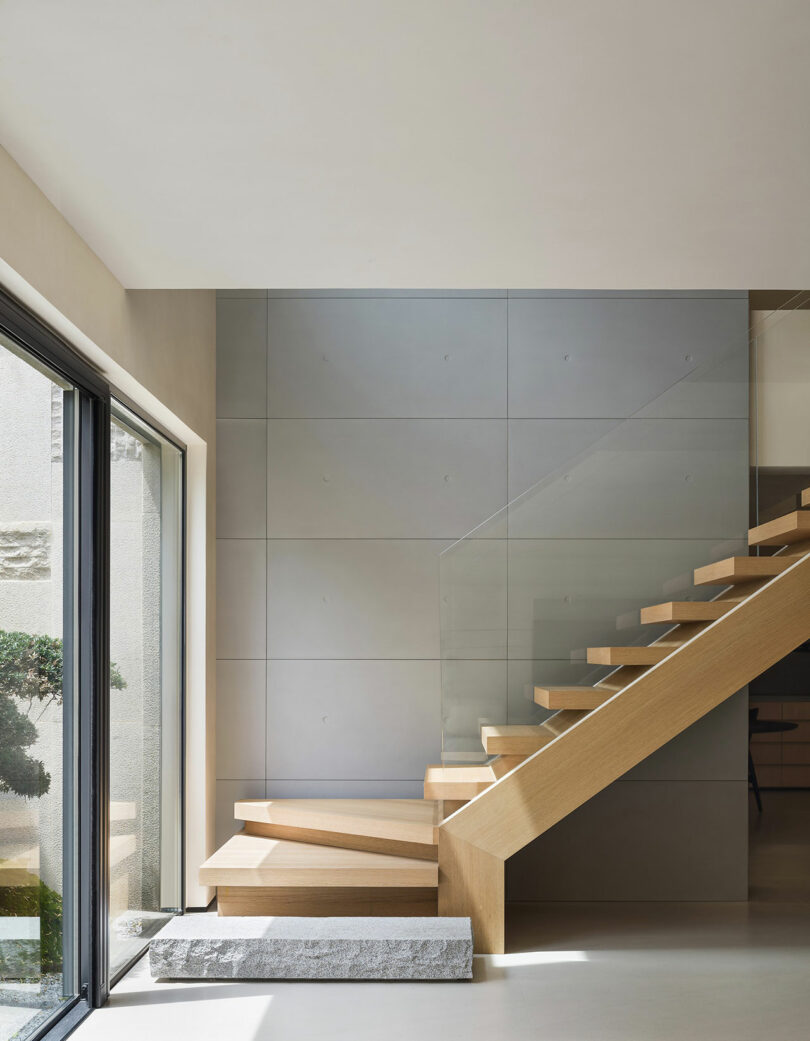 A modern interior with wooden floating stairs, gray panel walls, and large windows with a view of a courtyard.