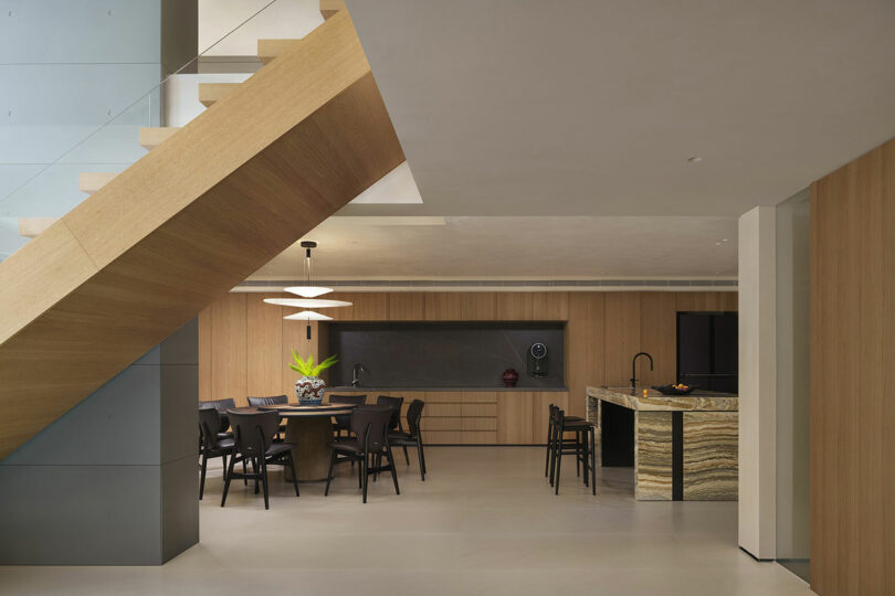 Modern interior with wooden staircase, open dining area featuring a round table with black chairs, and a kitchen with marble countertops and black fixtures. Warm wood tones and minimalistic design.