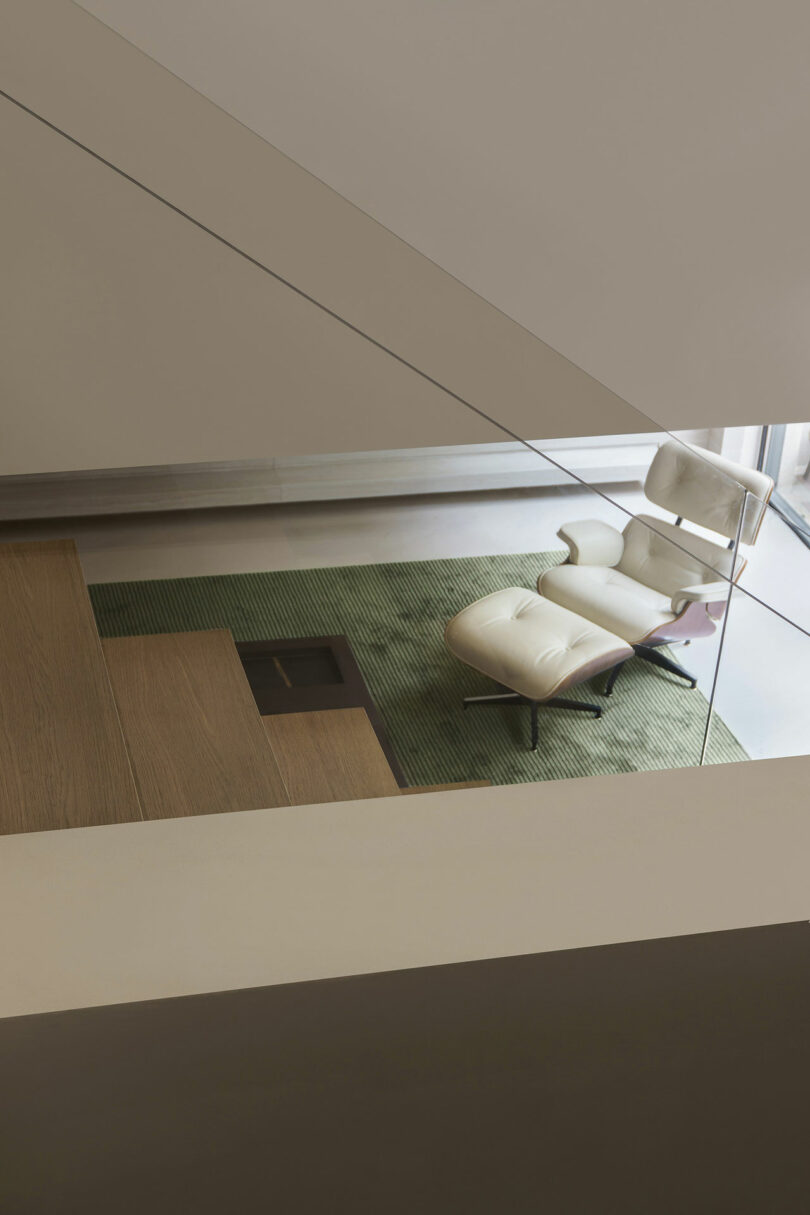 A beige upholstered lounge chair with an ottoman is set on a green rug near large windows, viewed from an overhead angle with a wooden staircase in the foreground.