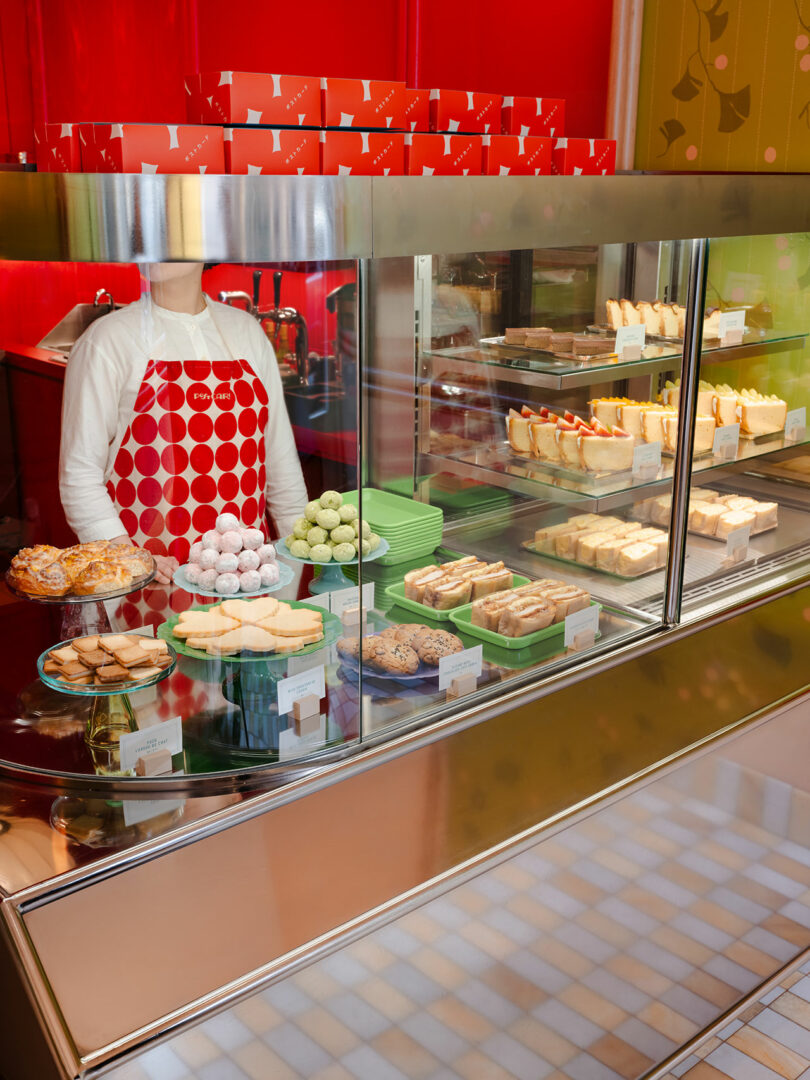 A person behind a bakery display counter filled with various pastries, cakes, and desserts. The person is wearing a red and white patterned apron and the background features red boxes.