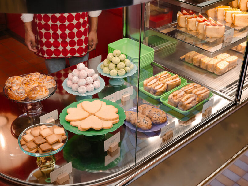A bakery display case featuring assorted pastries, cookies, and cakes. A person wearing a red polka dot apron stands behind the counter.
