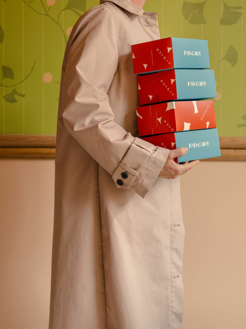 Person in a beige trench coat holding multiple boxed pastries against a green floral pattern background.