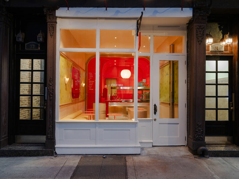 Nighttime view of a brightly lit cafe with red interior, large front window, white doors, and a hanging spherical lamp. Flanked by dark entrance doors on either side.