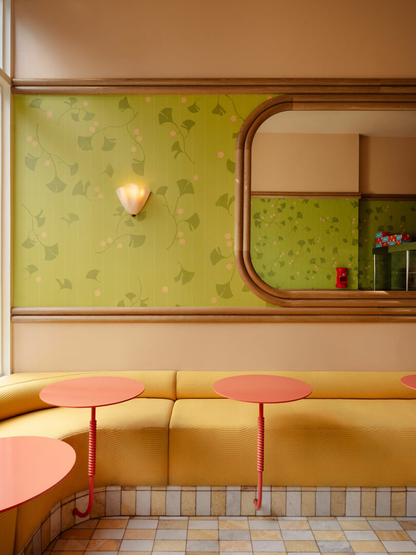 A brightly lit cafe interior with yellow seating, red circular tables, green floral wall patterns, and a large mirror reflecting the room.