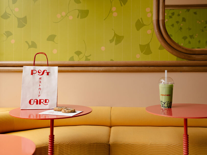 A small round table with a postcard-themed paper bag, cookies, and a bubble tea sits in a room with green patterned walls and yellow upholstered seating.