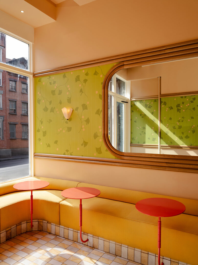 A brightly lit cafe interior with green patterned wallpaper, a large mirror, yellow cushioned seating, red round tables, tiled floor, and large windows showing a red brick building outside.