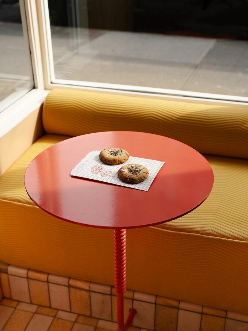 Two cookies on a patterned napkin rest on a small, round red table by a yellow bench with a window in the background.