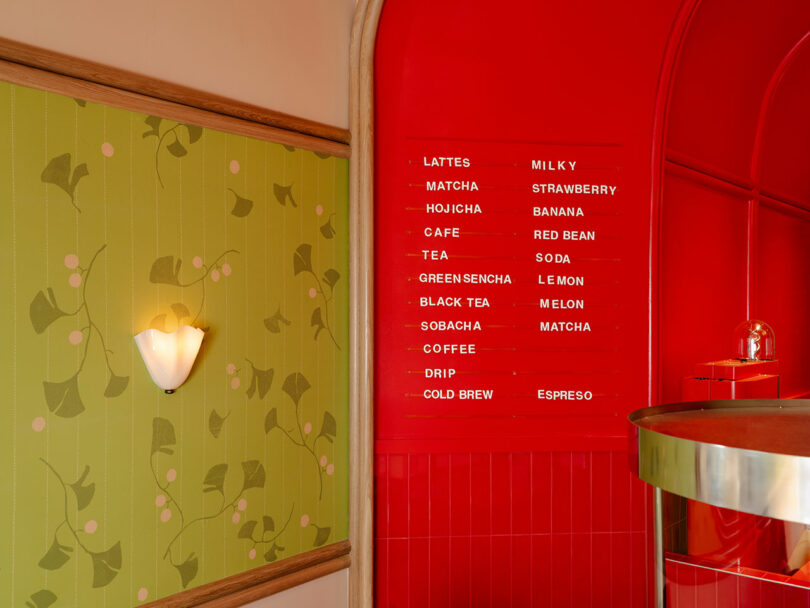 A menu mounted on a red wall lists beverages such as lattes, matcha, and hojicha alongside various flavors. The decor includes green wallpaper with a floral design and a wall light.