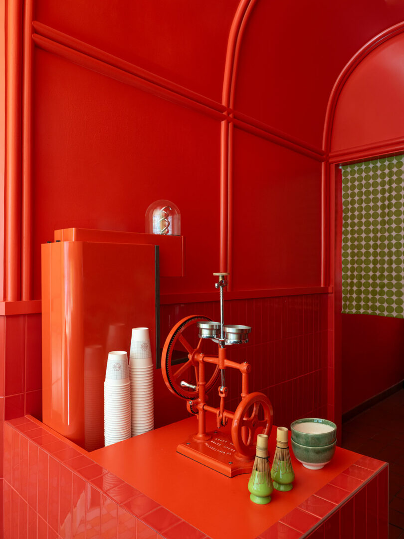 A minimalist red interior features an orange juice squeezer, stacked paper cups, and two matcha bowls with whisks on a red tiled counter.