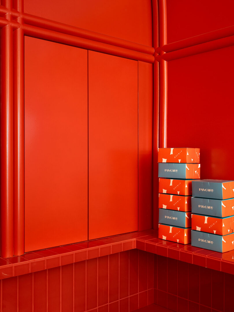 A stack of orange and gray Emporio boxes is placed on a red counter against a red wall with panels.