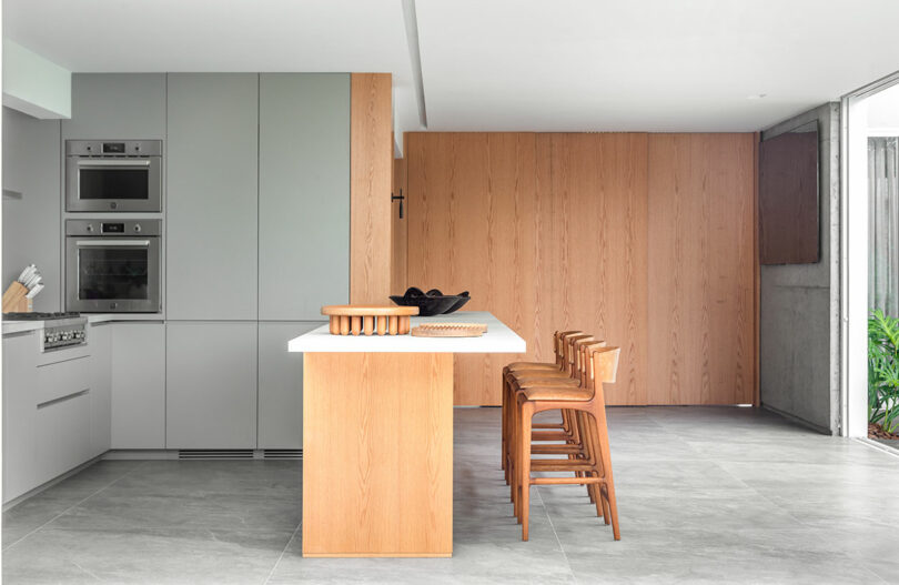 A modern kitchen with sleek gray cabinetry, built-in ovens, a wood-accented island with a white countertop, and four wooden bar stools on a grey tiled floor. Large windows provide natural light.