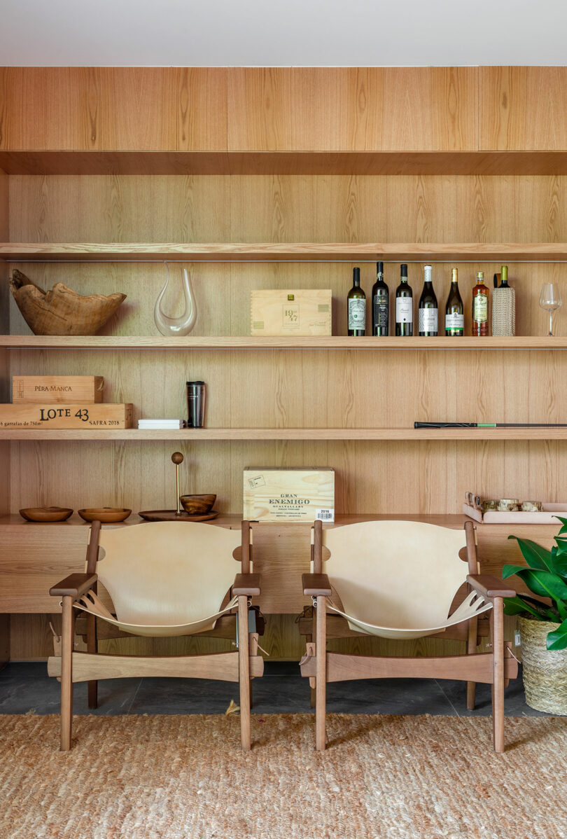 A wooden shelving unit with wine bottles, glasses, and decorative items behind two beige leather chairs on a textured rug, next to a green plant.