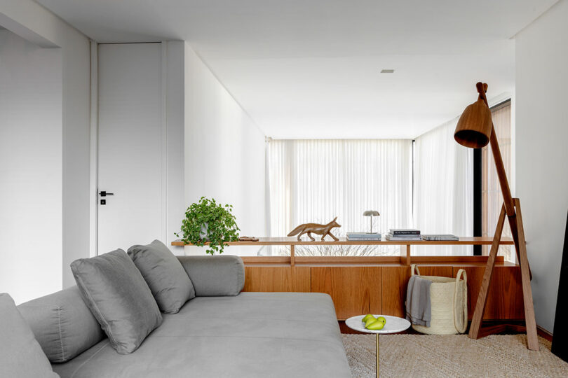 A modern living room with a gray sectional sofa, small round table with green apples, potted plant, and a wooden room divider. White curtains and a floor lamp are in the background.