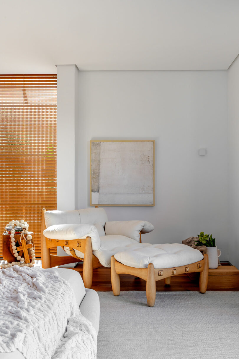 A modern living room with a wooden chair and ottoman with white cushions, a white textured blanket on a gray sofa, wooden blinds, a minimalist painting on the wall, and small plant on a wooden surface.