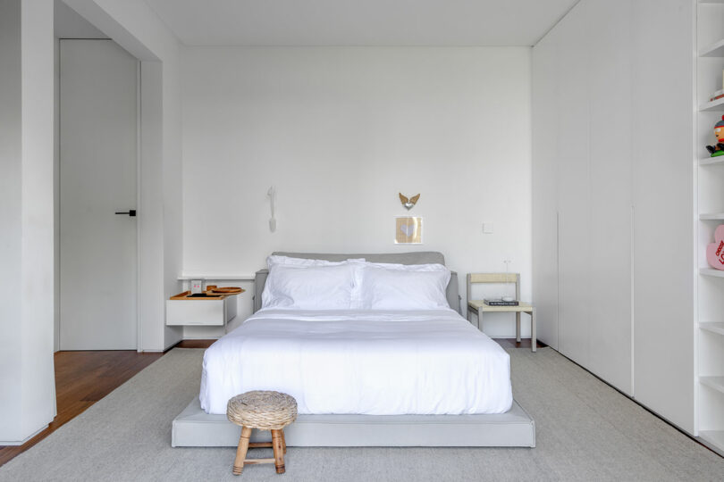 A minimalistic bedroom with a double bed, white bedding, a small stool at the foot, and two nightstands. The room features white walls, a closed door, and built-in shelves.