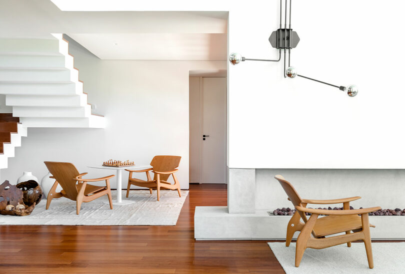 A modern living room features wooden chairs and a small table with a chess set. The room has a minimalist design with white walls, a staircase, and a unique light fixture.