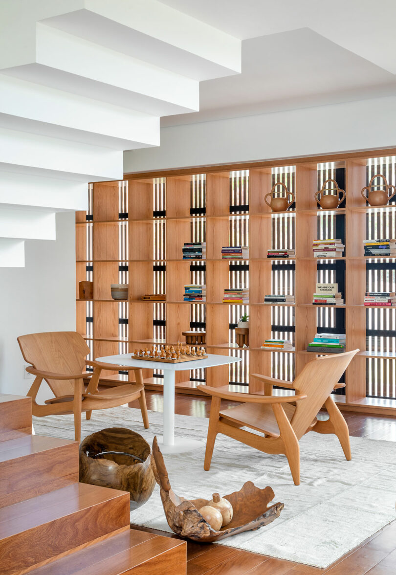 Modern living space featuring wooden bookshelves, books, and decorative items, with two wooden chairs facing a chessboard on a small table. White staircase and rug complete the minimalist design.