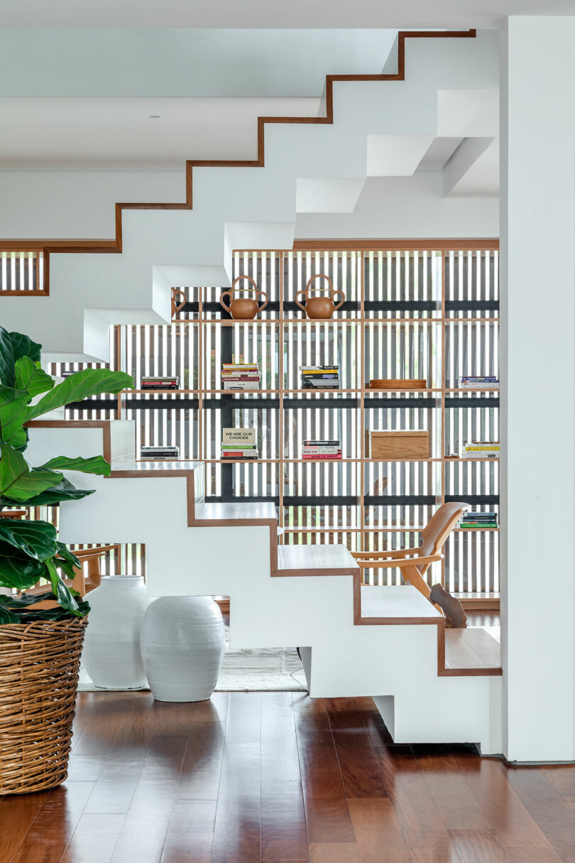 A modern interior with a white zigzag staircase, wooden accents, a plant in a woven basket, two white ceramic vases, and shelves with books and decor items along a slatted wall.