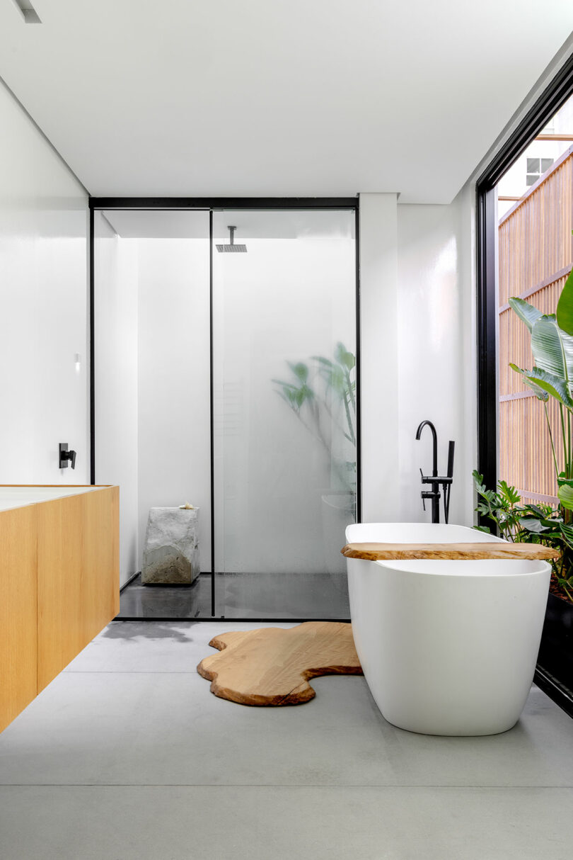 A modern bathroom featuring a white freestanding bathtub, wooden accents, a glass-enclosed shower, and large windows with potted plants.