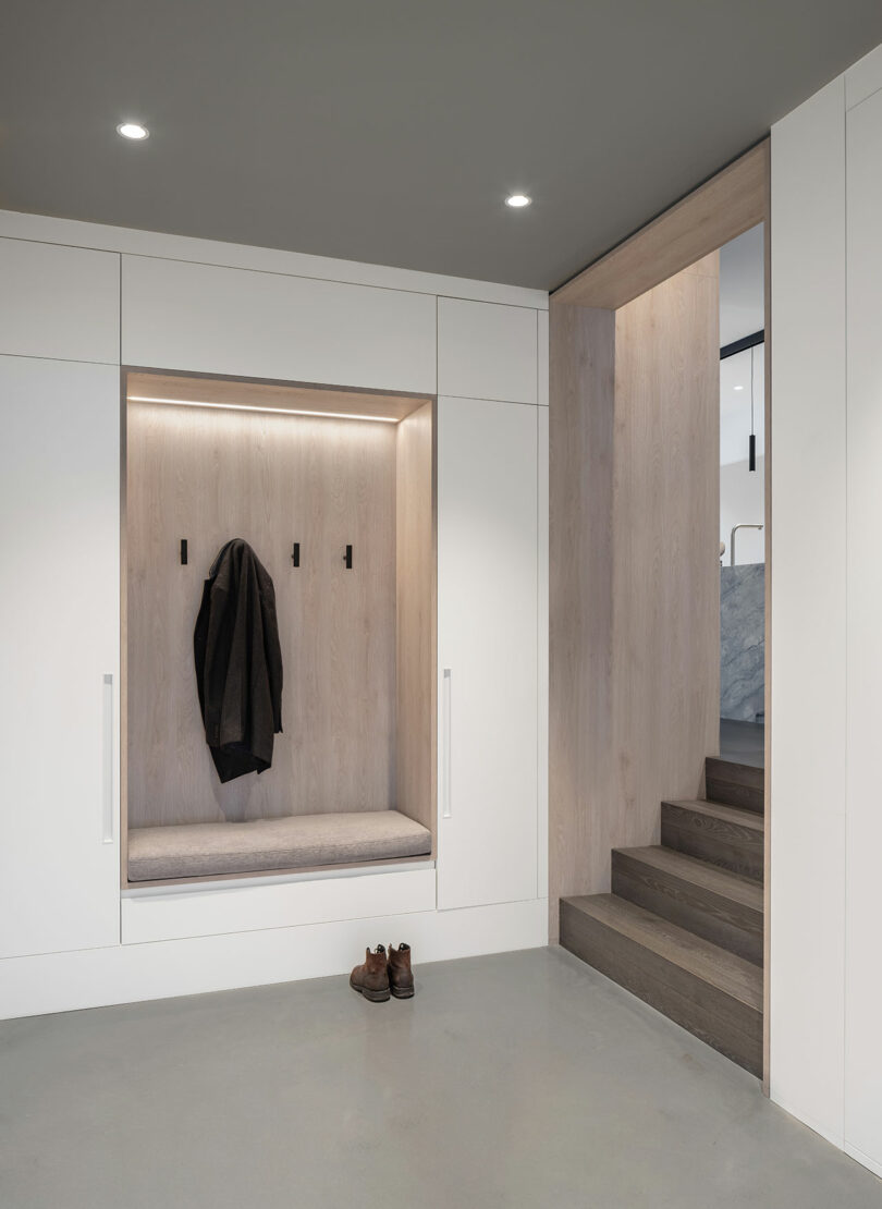 A modern entryway with built-in bench and coat hooks reflects sleek architecture, while a pair of shoes rests on the floor and stairs lead up to another room.