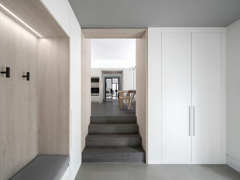 A modern interior with white walls and cabinets reflects contemporary architecture, featuring a built-in seating nook with hooks, and a view of the dining area through a doorway and steps.