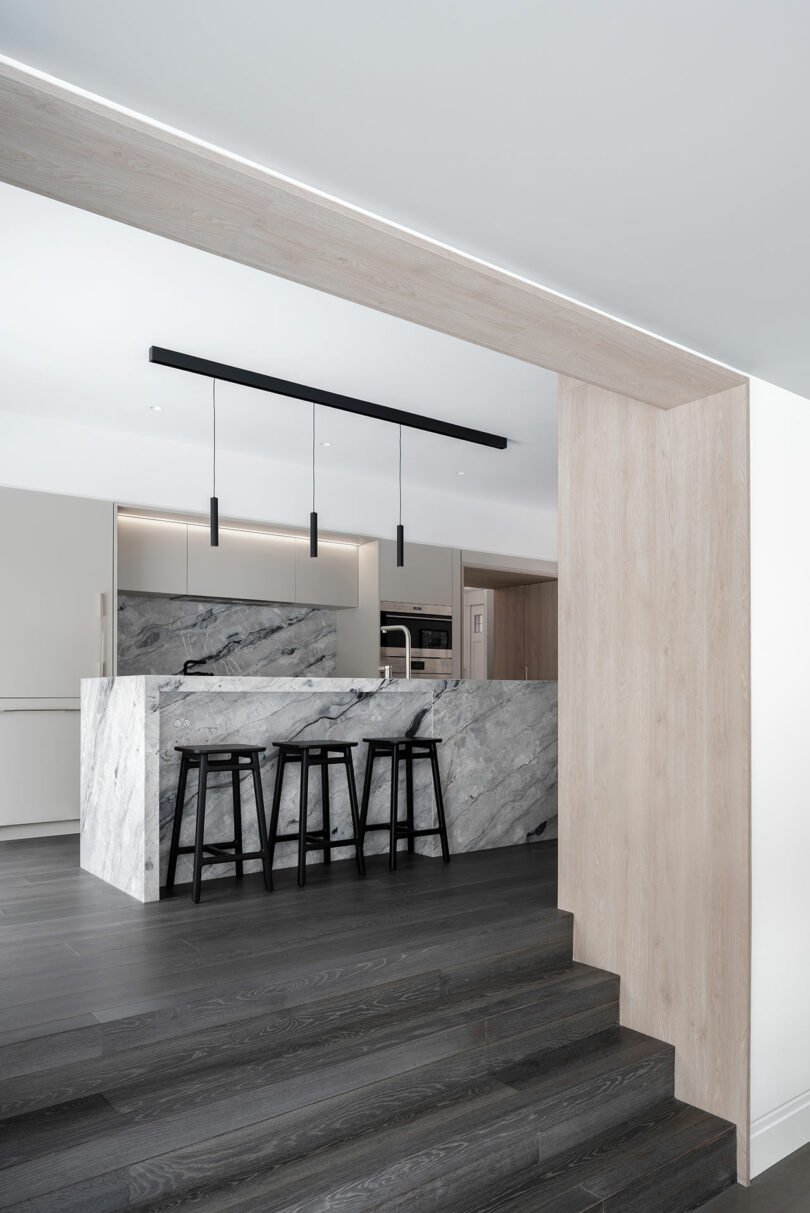 Reflecting contemporary architecture, this modern kitchen features a marble island countertop, three black bar stools, dark wood flooring, and minimalist pendant lights.