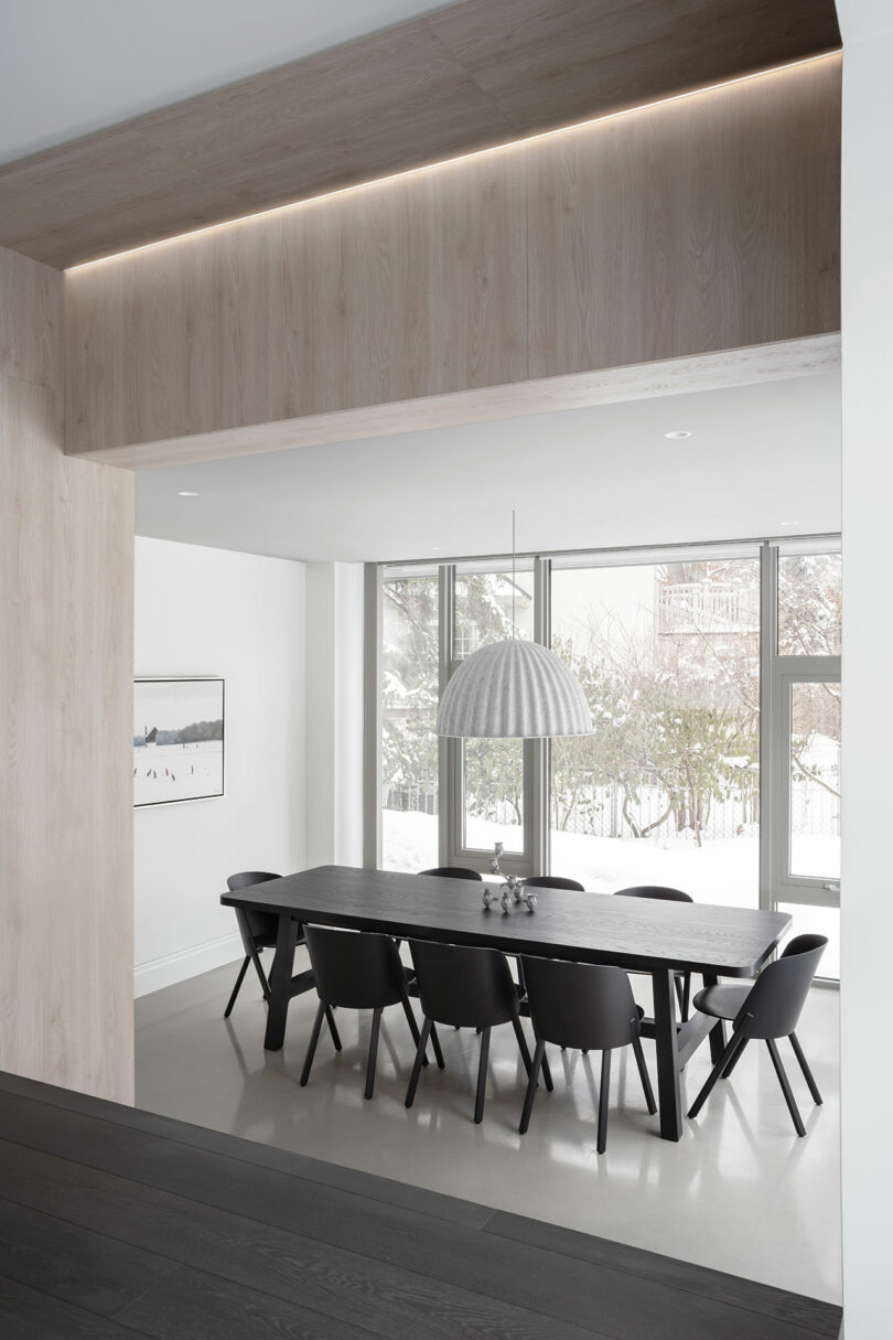 A modern dining room reflects sleek architecture, featuring a long black table with eight matching chairs, a large hanging light fixture, and floor-to-ceiling windows. A framed photo hangs elegantly on the white wall to the left.