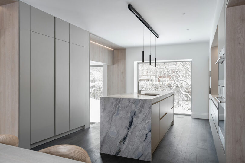A modern kitchen with light wood cabinets, a marble island, dark wood floors, and a large window that reflects the architecture of a snowy outdoor scene.