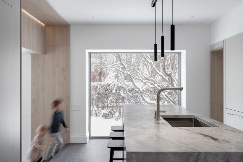 A modern kitchen with a marble island, black pendant lights, and a large window reflecting a snowy outdoor scene. Two children are blurred in motion near the cabinets, bringing life to the contemporary architecture.