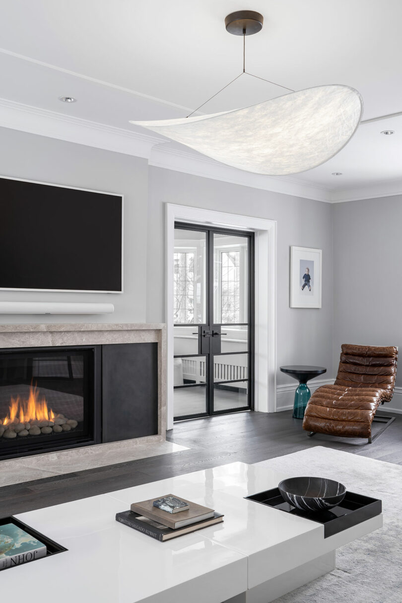 Modern living room with a lit fireplace, a wall-mounted TV, and a large curved light fixture. A sleek white coffee table and brown leather lounge chair reflect the architecture's sophistication. Large glass doors open to an exterior view.