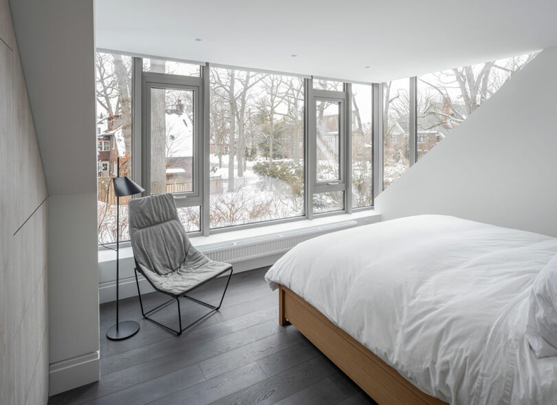 Modern bedroom with large windows, a white bed, and a gray chair with a floor lamp. The windows reflect the snowy outdoor landscape with trees and neighboring houses, creating an elegant blend of architecture and nature.
