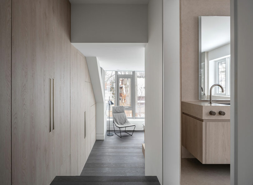 A minimalist hallway with light wood paneling reflects modern architecture as it leads to a cozy room featuring a gray rocking chair, large windows, and a bathroom with a sink and mirror on the right.