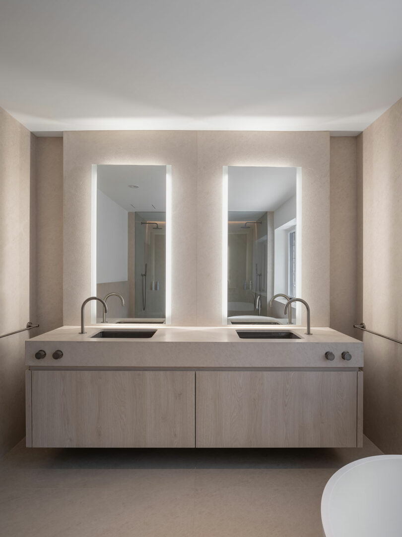Modern bathroom with a double vanity, backlit mirrors, and sleek faucets. The light-colored cabinetry and walls reflect architectural elegance, creating a clean and minimalist look.