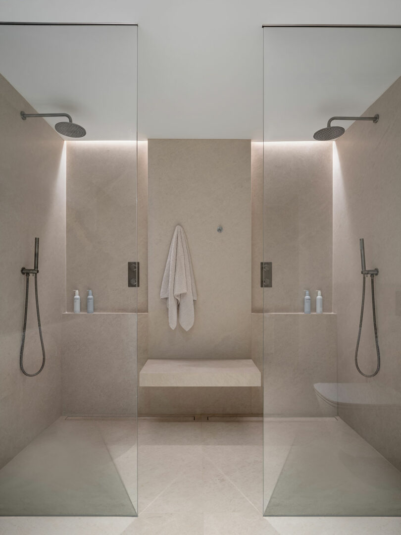 A modern shower room with two glass-enclosed shower stalls, each exemplifying Reflect Architecture’s signature style, equipped with a rainfall showerhead, handheld shower, and a niche with toiletries. A towel hangs on the wall between the stalls.