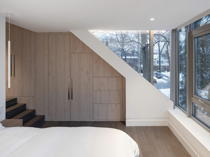 A modern bedroom featuring a custom wooden wardrobe with sleek black handles, large windows that reflect the stunning architecture of snowy trees outside, and a staircase leading to a lofted area.