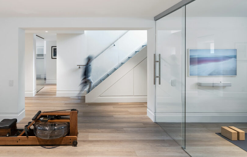 Modern home interior with a wooden rowing machine in the foreground, glass-walled room on the right reflecting architecture details, and a blurred figure descending a staircase in the background.