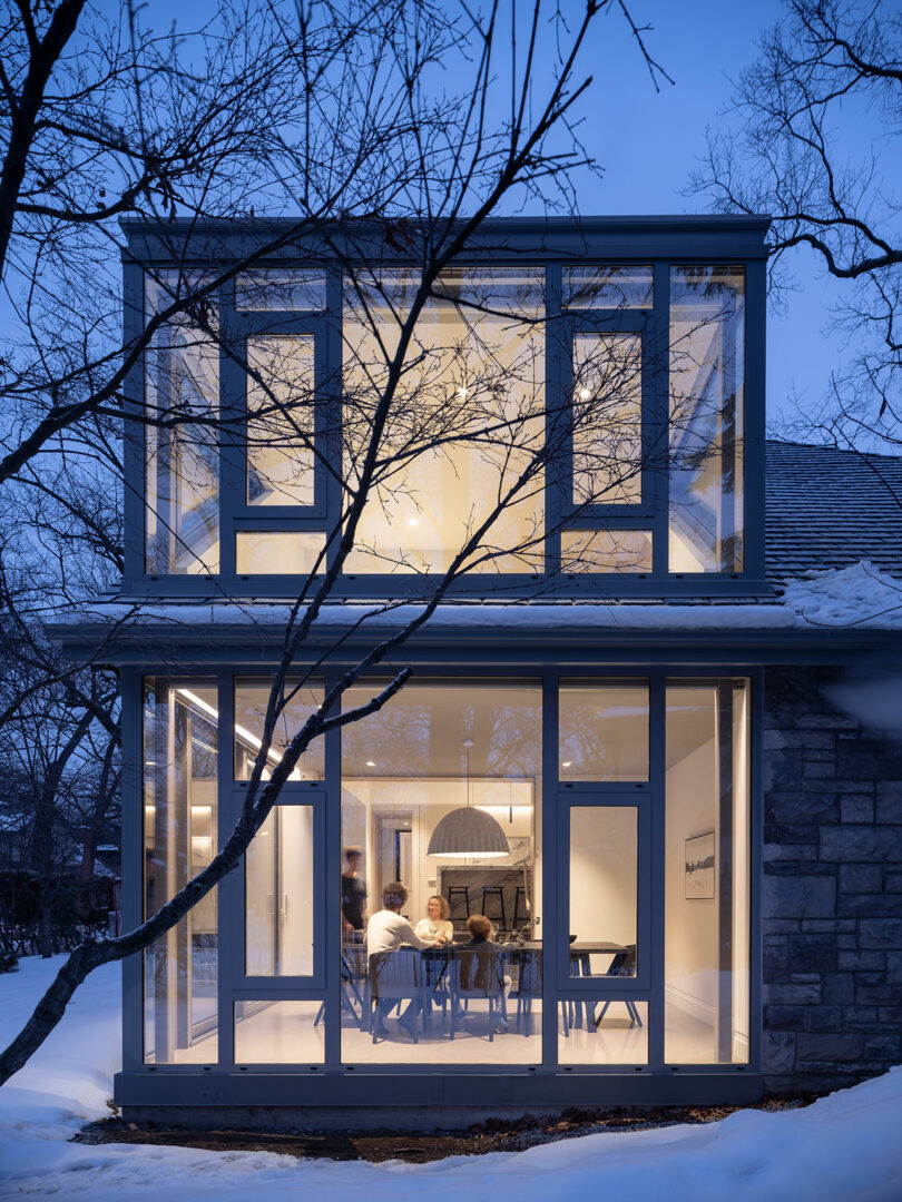 A modern glass house with two stories reflects the elegant architecture, illuminated from within during dusk. Snow covers the ground outside, and several people are seated around a table in the lower level.
