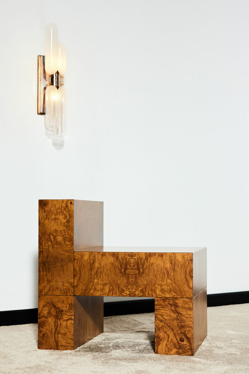 A wooden chair with a unique geometric design stands on a carpeted floor, positioned next to a modern wall sconce with three vertical frosted glass tubes.