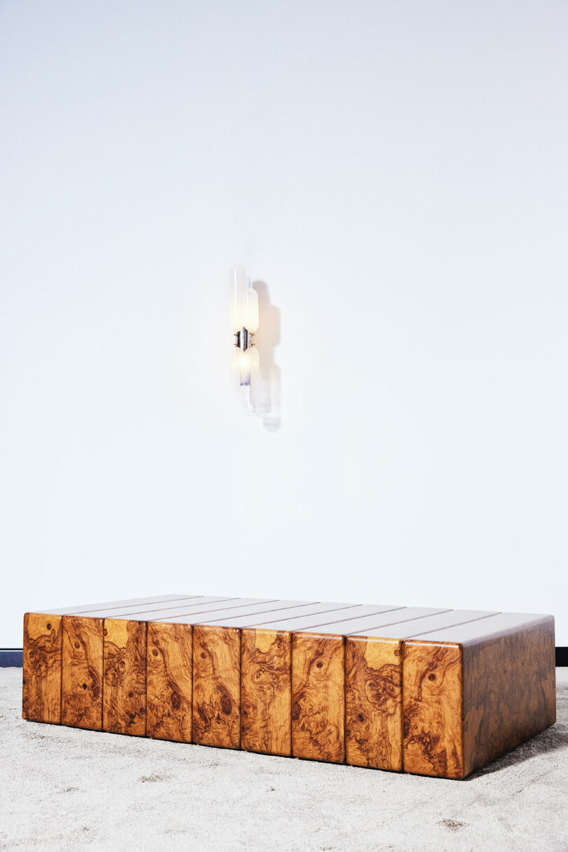 A rectangular wooden coffee table with a block design placed on a light-colored carpet in front of a white wall with a modern wall sconce above it.