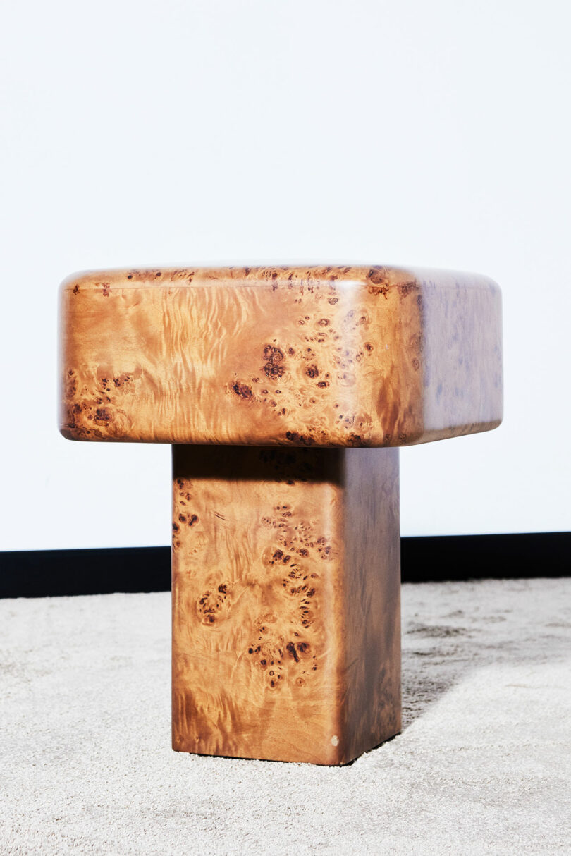 A modern rectangular wooden stool with a glossy finish, featuring a thick top and a sturdy rectangular base, placed on a light-colored carpeted floor against a white wall.