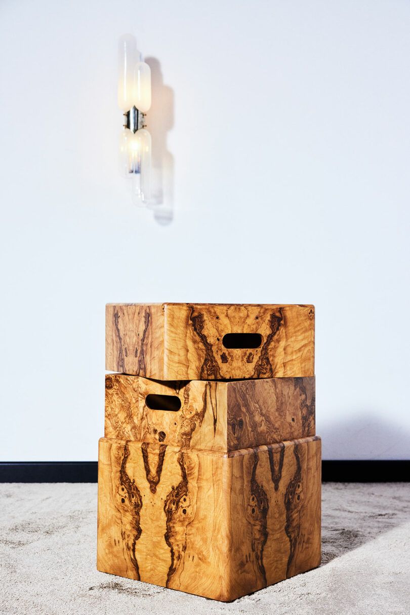 Two wooden storage boxes with cut-out handles are stacked on top of each other against a white wall. A modern wall sconce with two bulbs is mounted above them. The floor is carpeted.