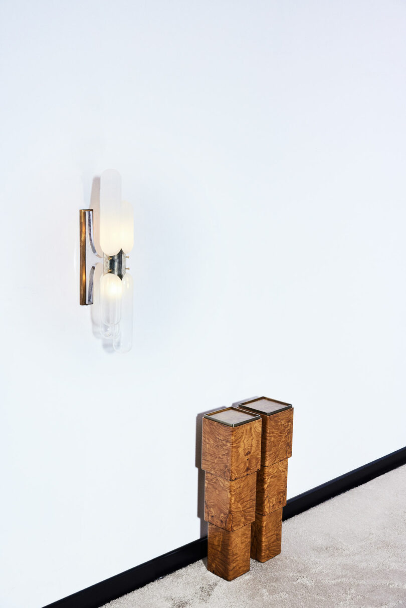 A contemporary wall light fixture is mounted on a white wall above a pair of stacked, rectangular wooden objects.