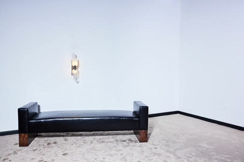 A minimalist room with a black leather bench positioned against a white wall. A modern wall sconce is illuminated above the bench. The floor is carpeted in a light beige color.