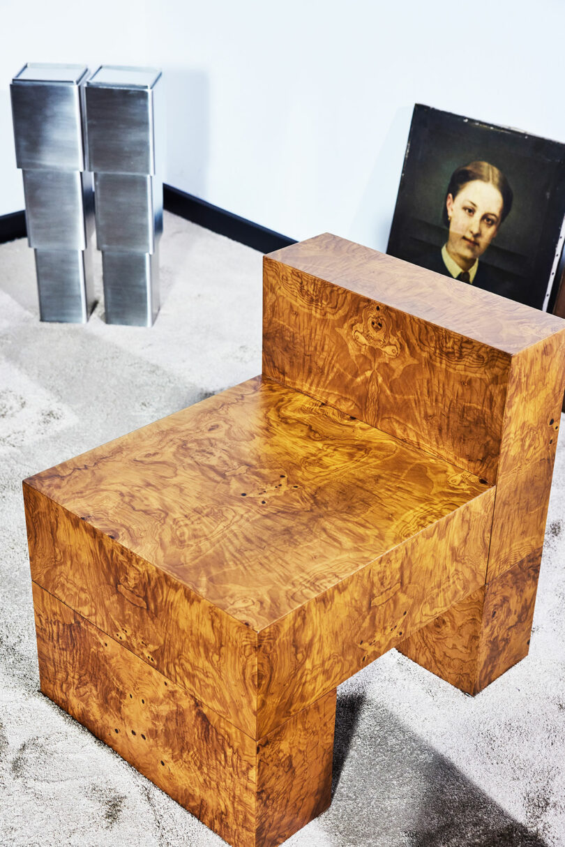 A cubic wooden chair with a natural finish sits on a beige carpet, next to two stacked metal boxes and a framed portrait leaning against a white wall.