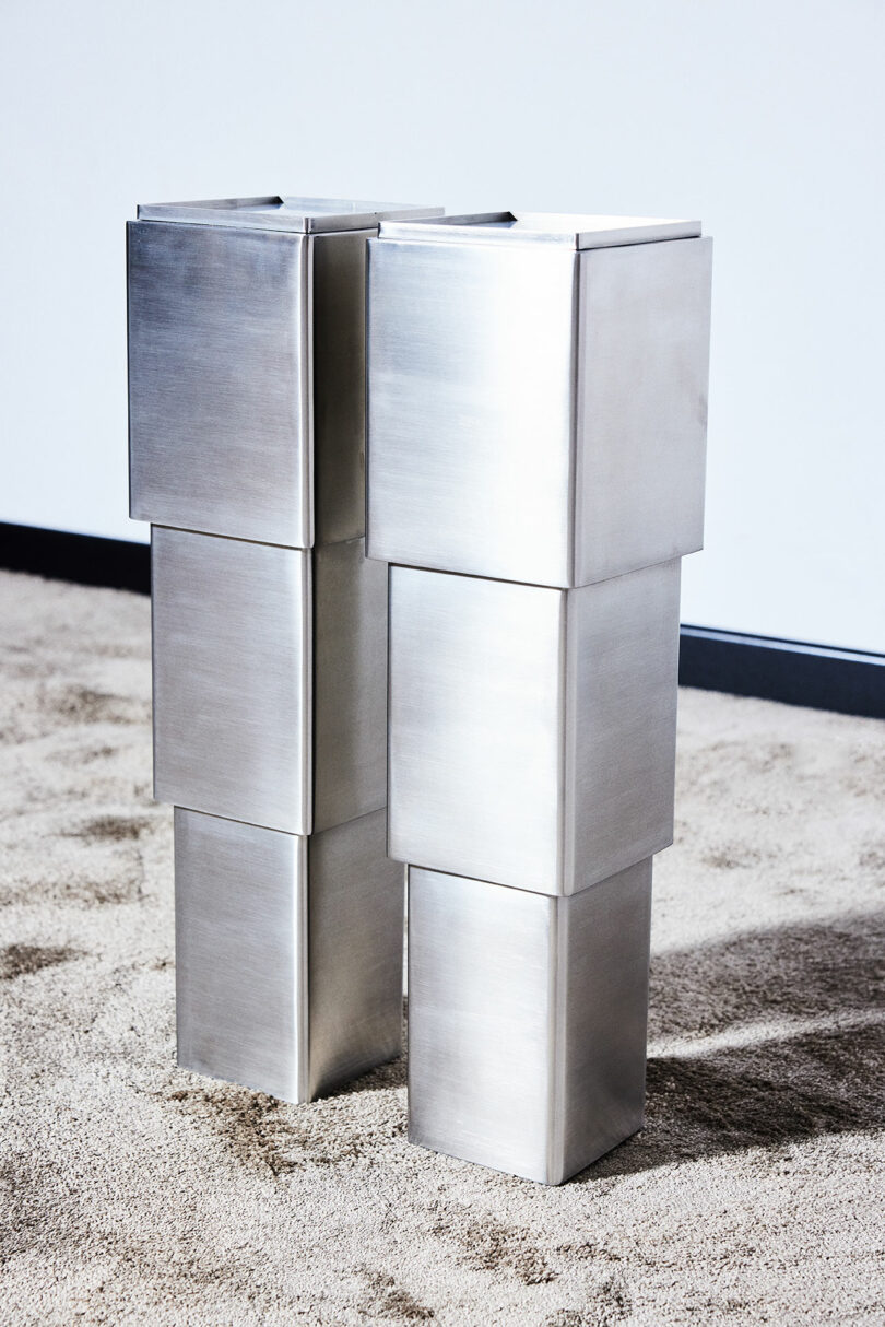 Two metallic, rectangular, minimalist sculptures are stacked vertically on a sandy surface against a white and black wall background.