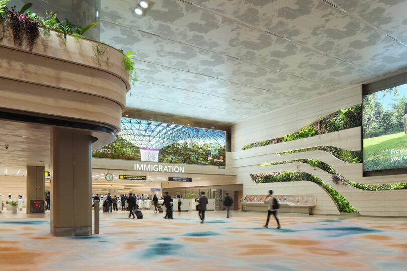 A spacious and modern airport terminal with an "Immigration" sign, numerous people walking, natural light from a skylight, and verdant wall decorations.