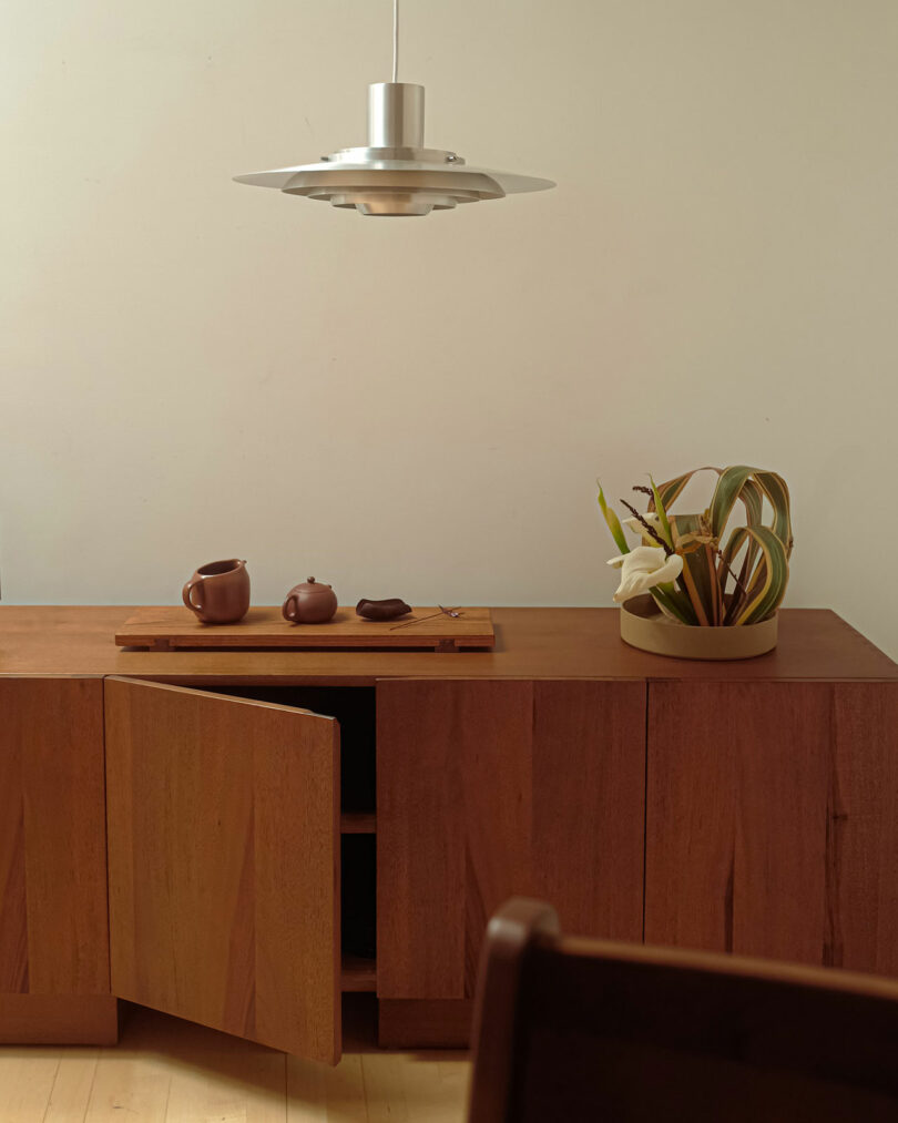 A wooden sideboard with one open door, topped with a tray holding a brown teapot and cups. Nearby, a beige pot with green plants is placed. A silver pendant light hangs above.