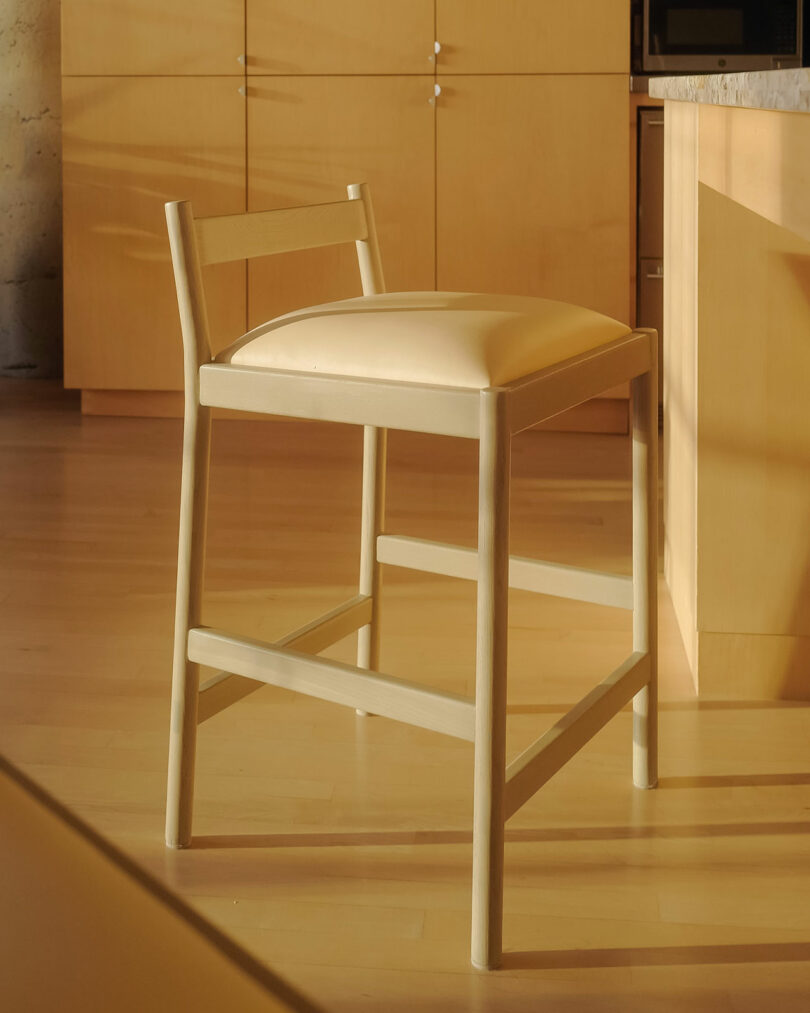 A light wooden chair with a cushioned seat is placed in a sunlit room with wooden cabinetry.