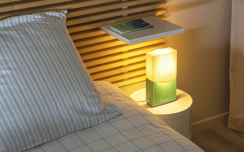 A bedside table with a glowing lamp, a book on a floating shelf, and a bed with a striped pillow and blanket in a modern bedroom